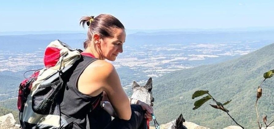 Woman sitting on a rocky ledge with a dog, overlooking a vast mountain landscape.